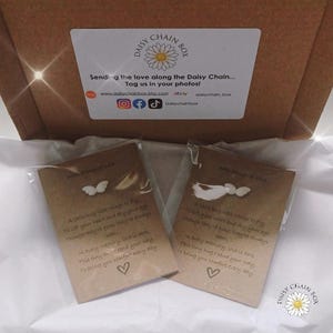 Può includere: Due piccoli ciondoli bianchi a forma di farfalla su biglietti con testo stampato. I biglietti sono all'interno di una scatola con l'etichetta "Daisy Chain Box". Il testo sui biglietti include una poesia sull'amore e il conforto.
