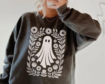 Può includere: Felpa grigio scuro con un disegno di fantasma bianco circondato da una ghirlanda floreale. Il fantasma ha un aspetto semplice e amichevole. La felpa è abbinata a leggings neri e un berretto bianco.