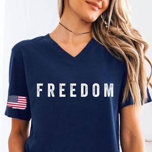 May include: Navy blue V-neck t-shirt with the word "FREEDOM" in white block letters. The American flag is on the left sleeve. The shirt is paired with denim shorts.