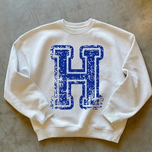May include: White crewneck sweatshirt with a large, distressed blue letter "H" printed on the front. The sweatshirt has long sleeves and a classic, casual style. The design gives a vintage, collegiate feel.