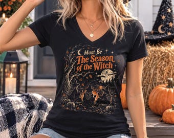 May include: A black V-neck t-shirt featuring a graphic print of three witches sitting around a campfire under a moonlit sky. The text reads, "Must Be The Season of the Witch." The design includes trees, falling leaves, and pumpkins in orange and gold.