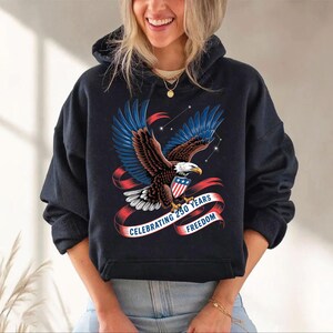 A woman wearing a black hoodie with a graphic design of an eagle and a red ribbon that says "Celebrating 250 years of freedom".