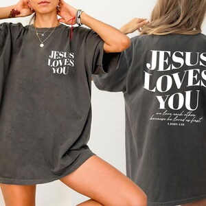 A person wearing a gray t-shirt with the text "JESUS LOVES YOU" printed on it, posing with their hands on their head.