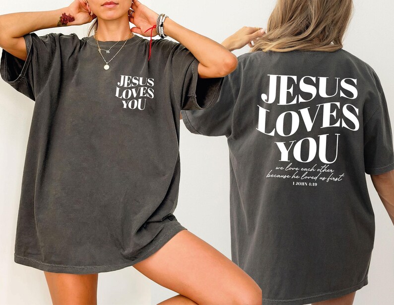 A person wearing a gray t-shirt with the text "JESUS LOVES YOU" printed on it, posing with their hands on their head.