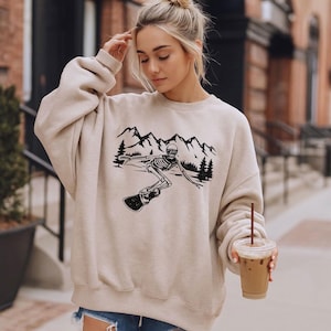 A young woman wearing a beige sweatshirt with a graphic of a skeleton riding a skateboard, holding a drink in her hand.