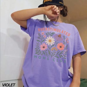 A person wearing a purple t-shirt with a floral design and text, standing in front of a white wall.