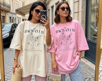 May include: Two oversized t-shirts, one cream and one pink, with the text "The Bach Club" and a graphic of a woman in a martini glass. The shirts also read "Emma's Bachelorette Party" and "Boston City 2023".