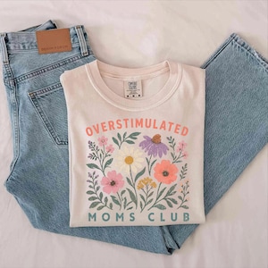 a white t-shirt with a floral design and the text "Oversaturated Moms Club" printed on it, along with a pair of blue jeans.
