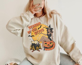 May include: Beige sweatshirt with a graphic that says "Season of the Witch". The graphic features a witch sitting on a pumpkin, a cauldron, bats, and a full moon. The person in the image is wearing a white baseball cap with the word "Karma" on it.