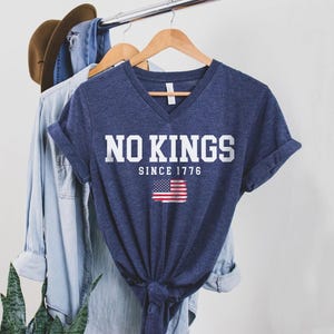A blue t-shirt with the text "NO KINGS SINCE 1776" printed on it, along with an American flag design, is hanging on a clothing rack.