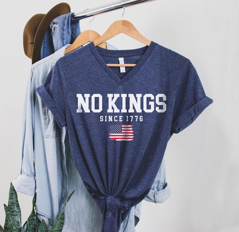 A blue t-shirt with the text "NO KINGS SINCE 1776" printed on it, along with an American flag design, is hanging on a clothing rack.