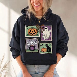 A woman wearing a black hoodie with a Halloween-themed design featuring a pumpkin, ghost, and other spooky elements.