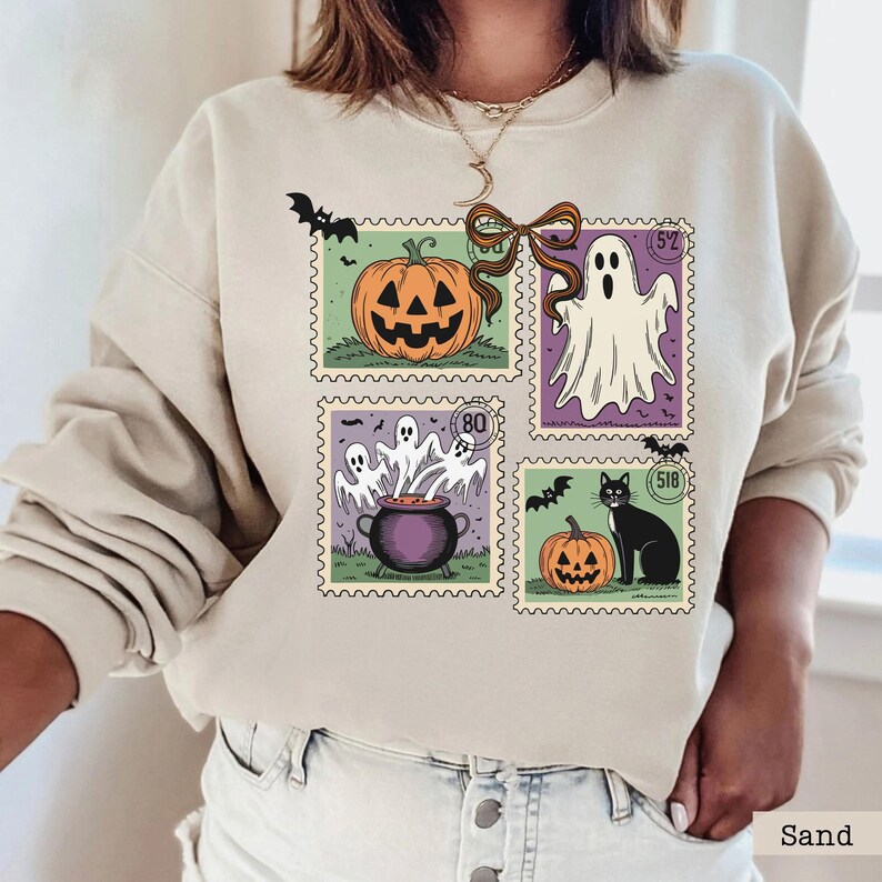 A woman wearing a white sweatshirt with a Halloween-themed design featuring a pumpkin, ghost, and bats.