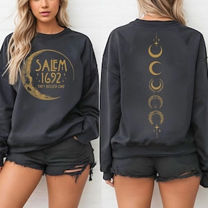 May include: Dark gray sweatshirt with gold text and a crescent moon design. The front reads "Salem 1692, They Missed One." The back features a vertical line of crescent moon phases. A casual, comfortable top.