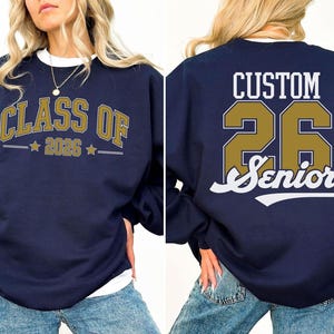A navy blue sweatshirt with gold text and numbers, reading "CLASS OF 2026" and "Custom 26 Senior" on the back.