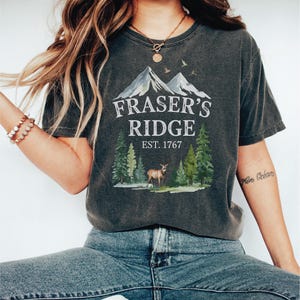 A woman wearing a gray t-shirt with a graphic design of a mountain and trees, along with the text "Fraser's Ridge Est. 1767".