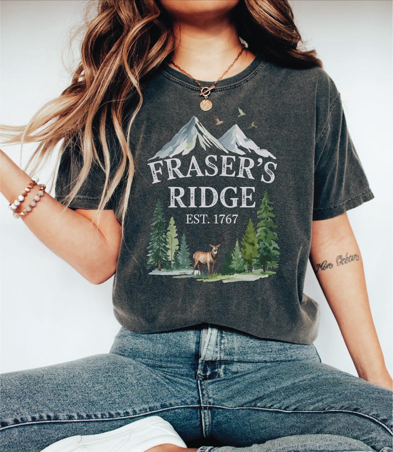 A woman wearing a gray t-shirt with a graphic design of a mountain and trees, along with the text "Fraser's Ridge Est. 1767".
