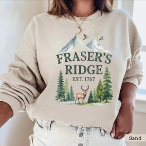 A woman wearing a beige sweatshirt with a graphic design featuring a mountain landscape, a deer, and the text "Fraser's Ridge Est. 1767".