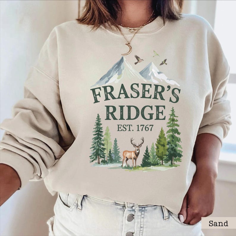 A woman wearing a beige sweatshirt with a graphic design featuring a mountain landscape, a deer, and the text "Fraser's Ridge Est. 1767".