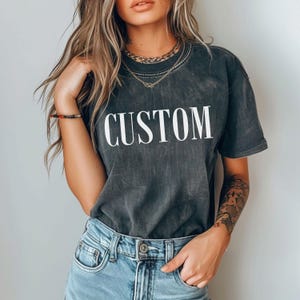 A woman wearing a black t-shirt with the word "CUSTOM" printed on it, paired with blue jeans and a necklace.