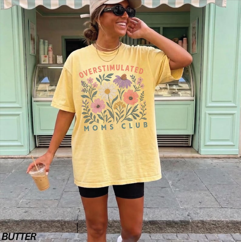 A woman wearing a yellow t-shirt with a floral design and sunglasses is standing in front of a storefront.