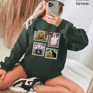 A person is sitting on the floor and taking a selfie with a smartphone, wearing a green sweatshirt with Halloween-themed designs.