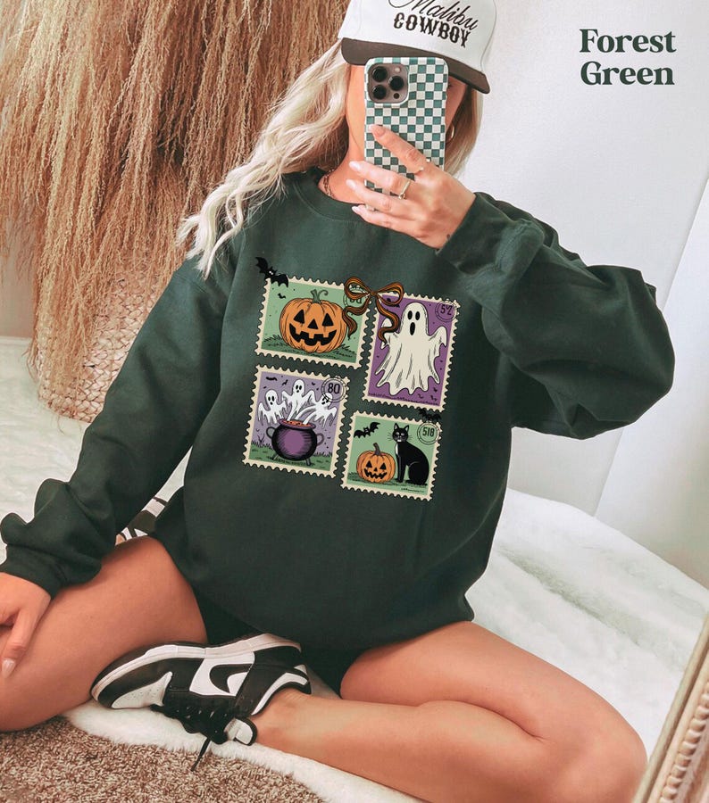 A person is sitting on the floor and taking a selfie with a smartphone, wearing a green sweatshirt with Halloween-themed designs.