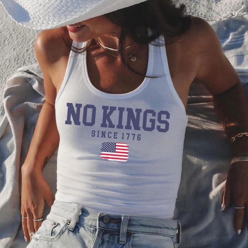 A woman wearing a white tank top with the text "NO KINGS SINCE 1776" and an American flag graphic, sitting on a blanket on the beach.