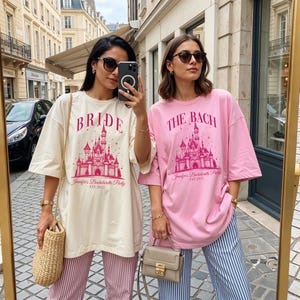 Two women wearing matching pink t-shirts with a castle design and the text "Bridal Jennifer's Bachelorette Party EST. 2005" are standing together and taking a selfie with a camera.