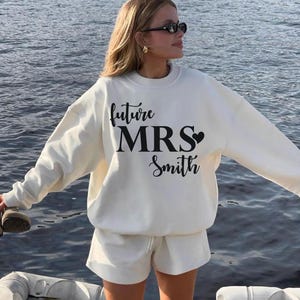 May include: Cream-colored sweatshirt and shorts set. The sweatshirt features the text "future MRS Smith" in black script with a heart. The model is wearing sunglasses and gold earrings.