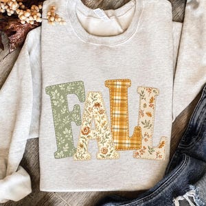 May include: A light gray crewneck sweatshirt featuring the word FALL printed in large block letters. Each letter has a unique pattern: the F is sage green with white leaves, the A is cream with orange flowers, the L is yellow plaid, and the L is cream with orange leaves.