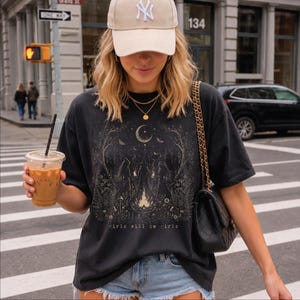 May include: A black t-shirt featuring a detailed graphic print of a crescent moon, forest elements, and two figures around a campfire, with the text "girls will be girls" printed below. The shirt is paired with a beige baseball cap and a black handbag.