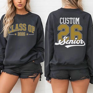 A woman wearing a black sweatshirt with the text "CLASS OF" and "26 Senior" printed on it, along with a number "26" and a letter "S" printed below.