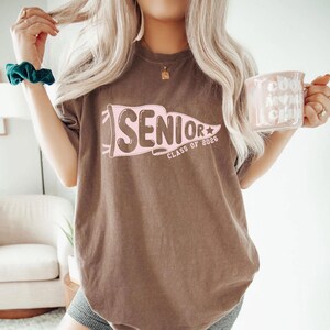 A woman with long blonde hair wearing a brown t-shirt with the text "SENIOR CLASS OF 2026" is holding a pink mug and a green scrunchie.