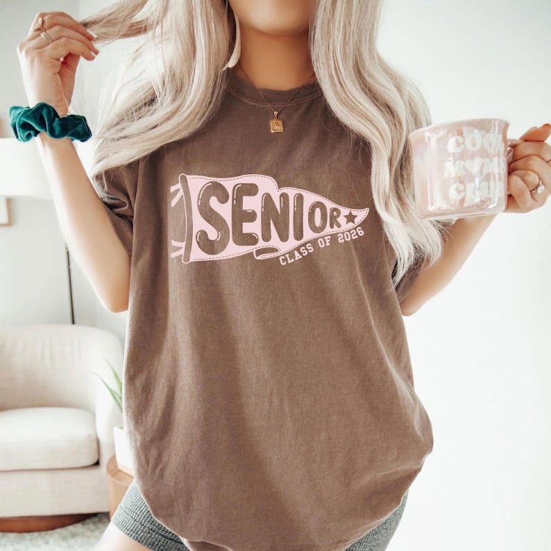 A woman with long blonde hair wearing a brown t-shirt with the text "SENIOR CLASS OF 2026" is holding a pink mug and a green scrunchie.