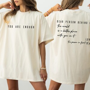 Two women wearing white t-shirts with the text "You are enough" and "Love. The person in front of you" printed on them.