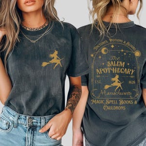 May include: Dark gray t-shirt featuring a gold witch silhouette on a broom. The back of the shirt displays the text "The Salem Apothecary" with additional text: "Tinctures, Potions & Elixirs", "Massachusetts", "Magic Spell Books & Cauldrons", and "EST. 1626".