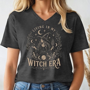 May include: A charcoal gray V-neck t-shirt featuring a beige graphic print of a person wearing a pointed hat, surrounded by celestial elements like moons, stars, and foliage. The text "LIVING IN MY WITCH ERA" is printed in a decorative font.