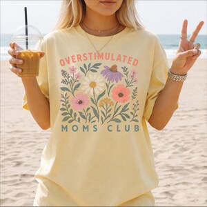 A woman wearing a yellow t-shirt with a floral design and text, holding a drink and making a peace sign gesture.