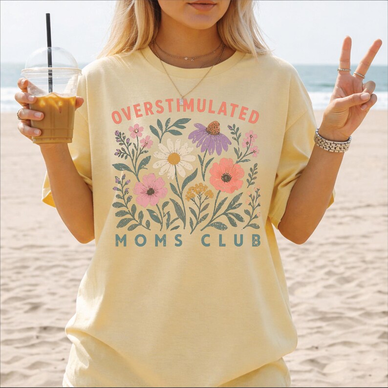 A woman wearing a yellow t-shirt with a floral design and text, holding a drink and making a peace sign gesture.