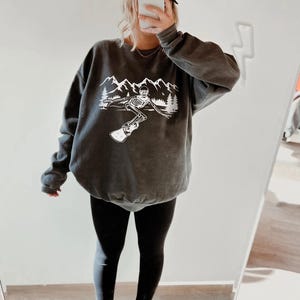 A person is taking a selfie in front of a mirror, wearing a gray sweatshirt with a graphic design and black leggings.