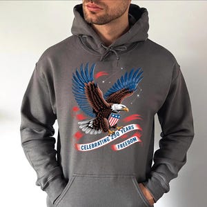 A man wearing a gray hoodie with a graphic design of an eagle and a red and blue ribbon banner that says "Celebrating 250 years of freedom".