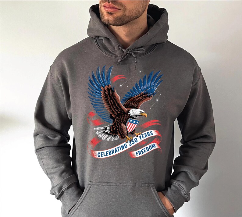 A man wearing a gray hoodie with a graphic design of an eagle and a red and blue ribbon banner that says "Celebrating 250 years of freedom".