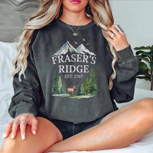 A woman wearing a gray sweatshirt with a graphic design of a mountain range and a deer, sitting on a bed.