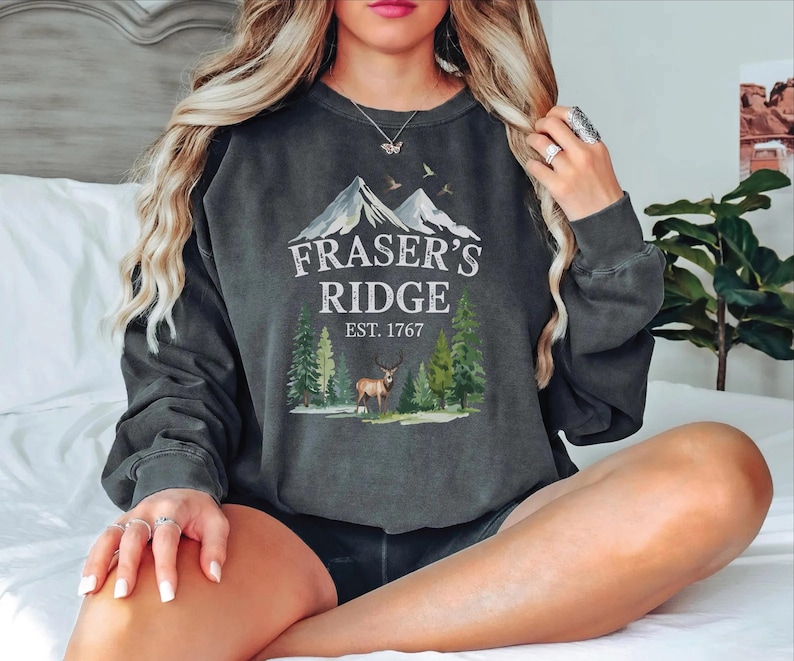 A woman wearing a gray sweatshirt with a graphic design of a mountain range and a deer, sitting on a bed.