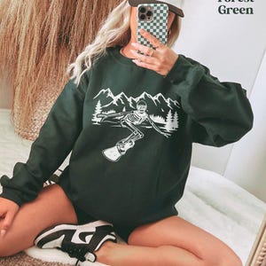 A person is taking a selfie in front of a mirror, wearing a green sweatshirt with a graphic design and a baseball cap.