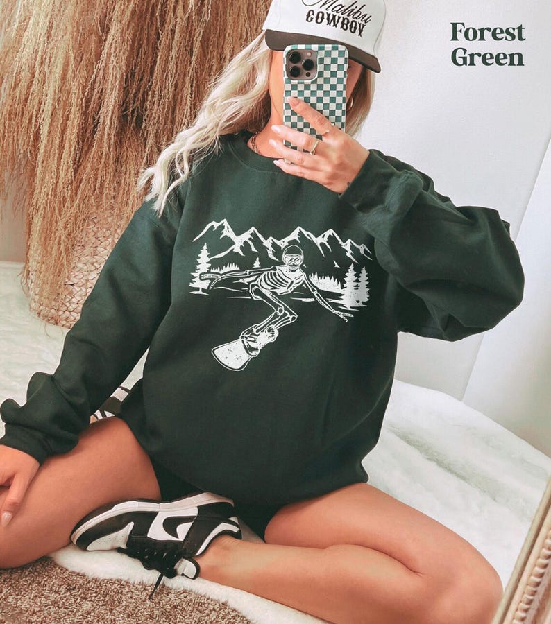 A person is taking a selfie in front of a mirror, wearing a green sweatshirt with a graphic design and a baseball cap.
