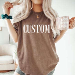 A woman with long blonde hair wearing a brown t-shirt with the word "CUSTOM" printed on it, holding a pink mug in her hand.