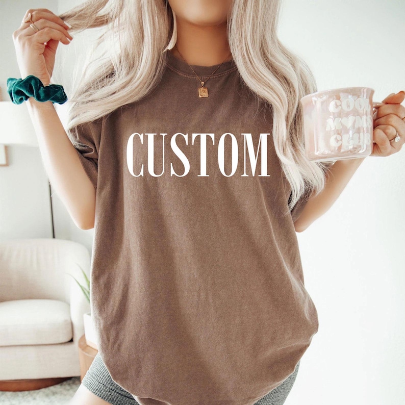 A woman with long blonde hair wearing a brown t-shirt with the word "CUSTOM" printed on it, holding a pink mug in her hand.