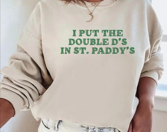 May include: A sand-colored sweatshirt with the text "I PUT THE DOUBLE D'S IN ST. PADDY'S" in green lettering. The sweatshirt has long sleeves and a crew neck. The bottom right corner of the image has the word "Sand".